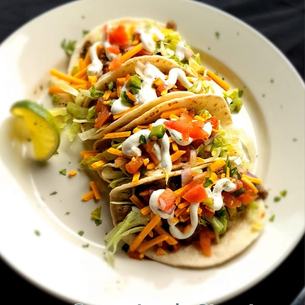 Three soft tacos filled with meat, cheese, lettuce, tomatoes, and sour cream, with a lime wedge.
