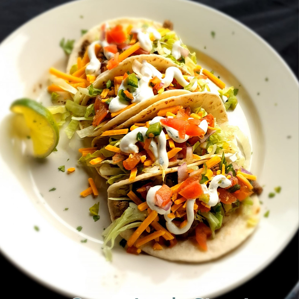 Three soft tacos filled with meat, cheese, lettuce, tomatoes, and sour cream, with a lime wedge.
